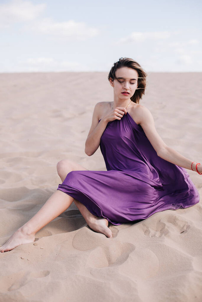 A young, slender girl in a beige dress with purple cloth in her hands posing in the desert in the wind - Zdjęcie, obraz