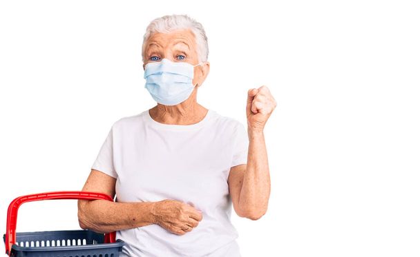 Senior beautiful woman with blue eyes and grey hair wearing shopping basket and medical mask screaming proud, celebrating victory and success very excited with raised arms  - Foto, Imagem