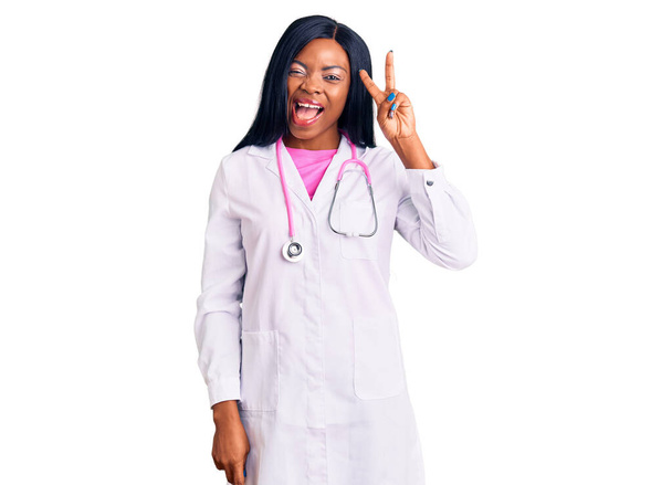 Mladá afro-americká žena v lékařském stetoskopu, usmívající se se šťastnou tváří, mrkající na kameru a mžourající prsty na znamení vítězství. číslo dvě.  - Fotografie, Obrázek