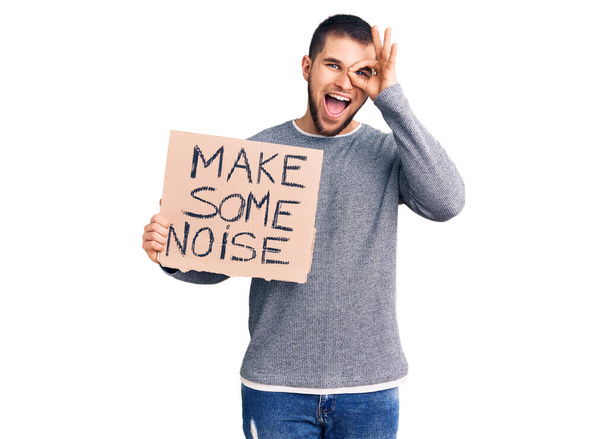 Young handsome man holding make some noise banner smiling happy doing ok sign with hand on eye looking through fingers  - Photo, Image