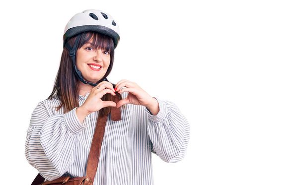 Jonge plus size vrouw met fietshelm en leren tas lachend in liefde met hartsymbool en vorm met handen. romantisch concept.  - Foto, afbeelding