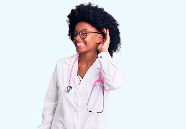 A fiatal afro-amerikai nő orvosi köpenyt és sztetoszkópot visel, és fültől fülig mosolyog, miközben a pletykákat és pletykákat hallgatja. süketség fogalma.  - Fotó, kép