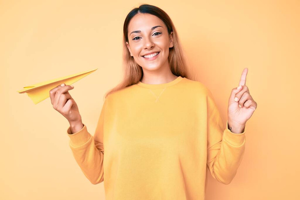 Young brunette woman holding paper airplane smiling happy pointing with hand and finger to the side  - 写真・画像