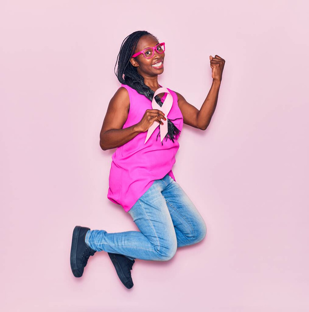 Jovem mulher afro-americana bonita usando óculos sorrindo feliz. Saltando com sorriso no rosto segurando fita de câncer rosa celebrando com punho para cima sobre fundo isolado - Foto, Imagem