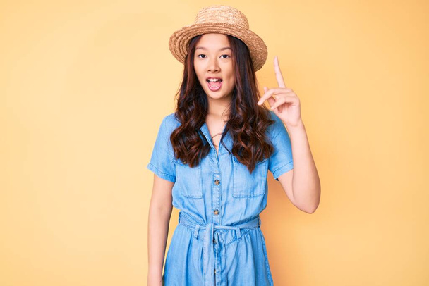 Junge schöne chinesische Mädchen mit Sommermütze zeigt mit erhobenem Zeigefinger auf erfolgreiche Idee. aufgeregt und glücklich. Nummer eins.  - Foto, Bild