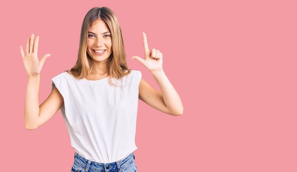 Schöne kaukasische Frau mit blonden Haaren trägt legeres weißes T-Shirt und zeigt mit Finger Nummer sieben nach oben, während sie selbstbewusst und glücklich lächelt.  - Foto, Bild