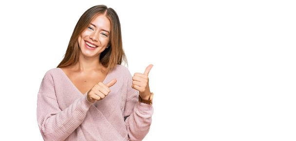 Beautiful blonde woman wearing casual winter pink sweater pointing to the back behind with hand and thumbs up, smiling confident  - 写真・画像