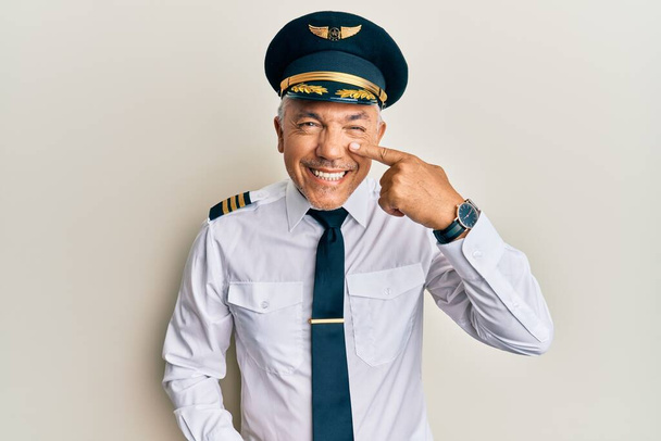 Hombre maduro guapo de mediana edad con uniforme de piloto de avión apuntando con el dedo a la mano y la nariz, sonriendo alegre. concepto de belleza  - Foto, Imagen