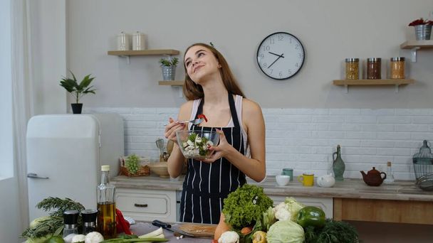 gelukkig jong vrouw eten vers rauw groente salade poseren in keuken met positieve emotie - Foto, afbeelding