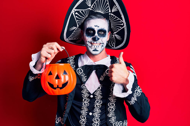 Young man wearing mexican day of the dead costume holding pumpkin smiling happy and positive, thumb up doing excellent and approval sign  - Photo, Image