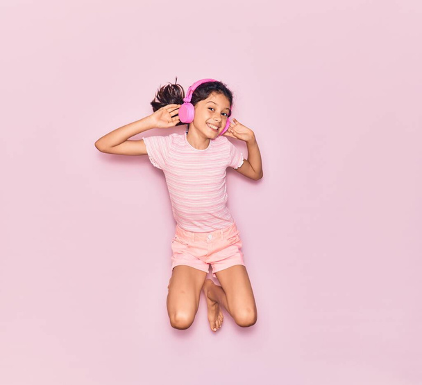 Adorável criança hispânica menina sorrindo feliz. Saltando com sorriso no rosto ouvindo música usando fones de ouvido rosa sobre fundo isolado - Foto, Imagem
