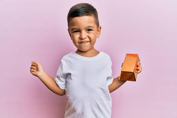 Adorable latin toddler holding gold ingot smiling happy pointing with hand and finger to the side  - Fotoğraf, Görsel