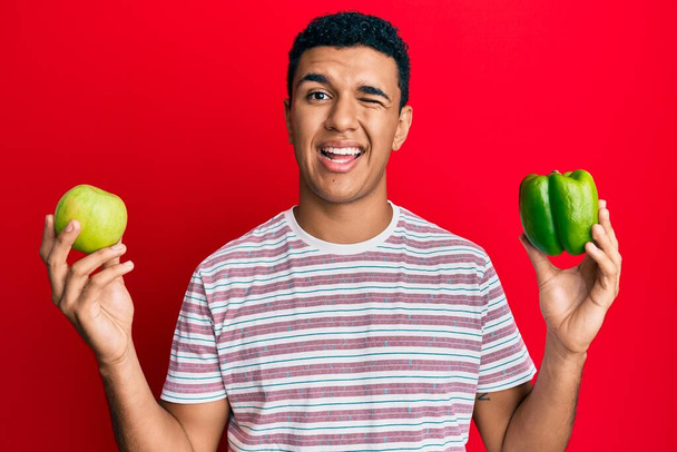 Young arab man holding green apple and pepper winking looking at the camera with sexy expression, cheerful and happy face.  - Foto, Imagem