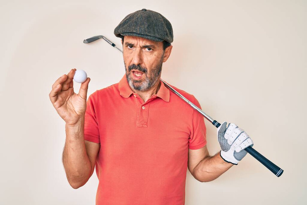 Uomo ispanico di mezza età in possesso di golf club e palla in faccia shock, cercando scettico e sarcastico, sorpreso a bocca aperta  - Foto, immagini