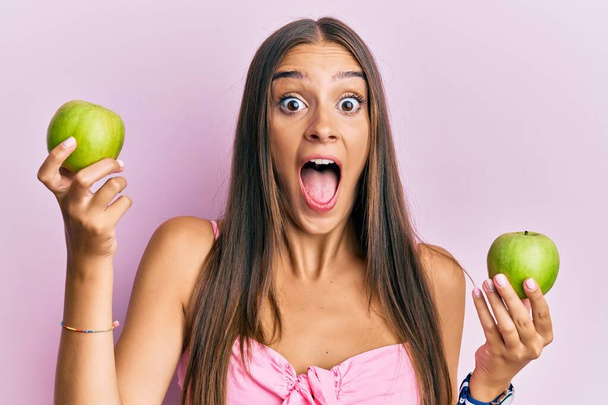 Młoda Latynoska kobieta trzyma zielone jabłka świętując szaleństwo i zdumiony sukcesem z otwartymi oczami krzycząc podekscytowany.  - Zdjęcie, obraz