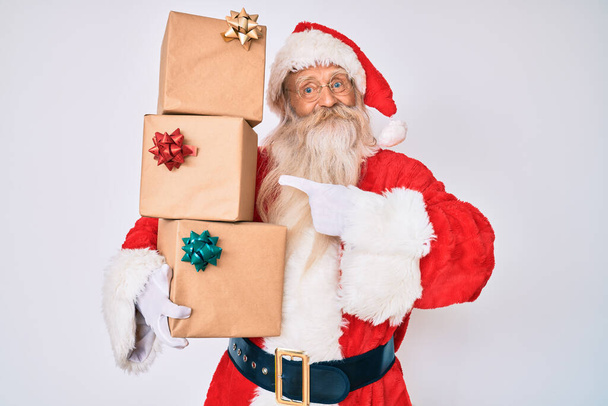Gri saçlı, uzun sakallı, Noel Baba kostümü giyen yaşlı bir adam elinde hediyelerle mutlu bir şekilde elini ve parmağını göstererek gülümsüyor.  - Fotoğraf, Görsel