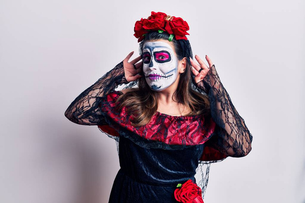 Young woman wearing day of the dead costume over white trying to hear both hands on ear gesture, curious for gossip. hearing problem, deaf  - Photo, Image