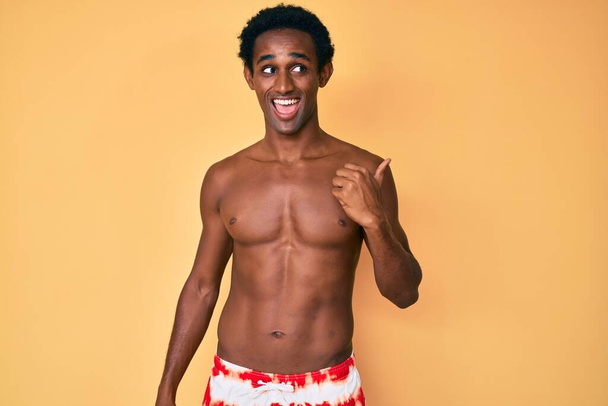 Africano guapo hombre usando traje de baño sonriendo con la cara feliz mirando y señalando a un lado con el pulgar hacia arriba.  - Foto, imagen