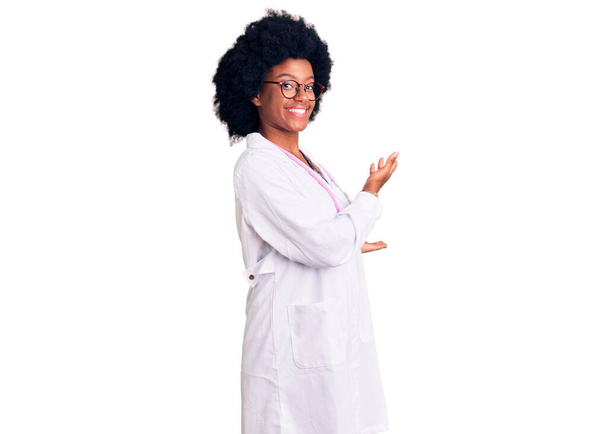 Молодая африканская американка в медицинском пальто и стетоскопе приглашает войти улыбающейся натуральной с открытой рукой  - Фото, изображение