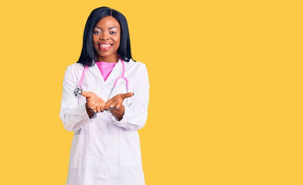 Doktor steteskop takan genç Afro-Amerikalı kadın avuçlarıyla gülümsüyor ya da el hareketi yapıyor. Bekleme ve koruma  - Fotoğraf, Görsel