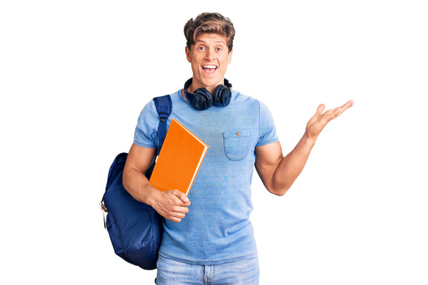 Fiatal jóképű férfi visel diák hátizsák és fejhallgató gazdaság könyv ünneplő győzelem boldog mosollyal és győztes kifejezés emelt kézzel  - Fotó, kép