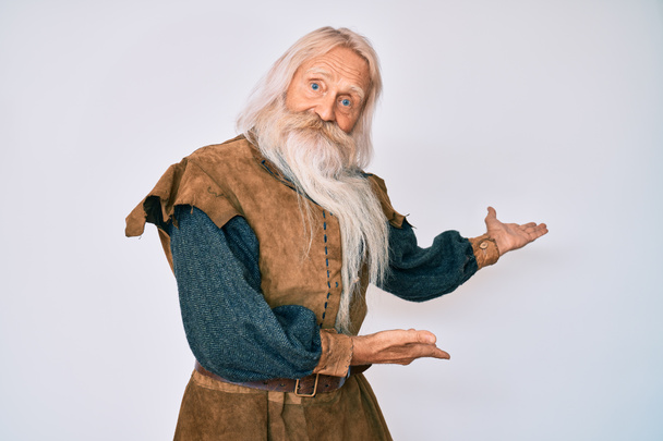 Vieil homme âgé aux cheveux gris et à la barbe longue portant un costume traditionnel viking invitant à entrer souriant naturel à la main ouverte  - Photo, image