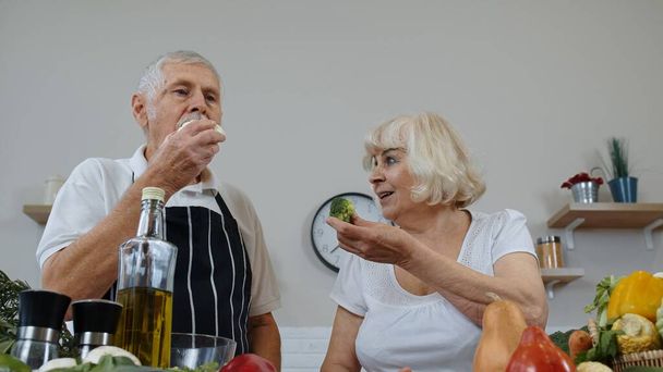 キッチンのシニアカップル。高齢者の祖母と祖父は生のブロッコリーとカリフラワーを食べる - 写真・画像