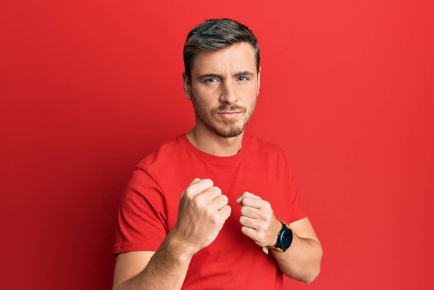 Handsome caucasian man wearing casual red tshirt ready to fight with fist defense gesture, angry and upset face, afraid of problem  - Photo, Image