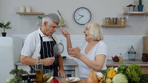 Feliz pareja de ancianos bailando mientras cocinan juntos en la cocina con verduras y frutas frescas - Foto, imagen