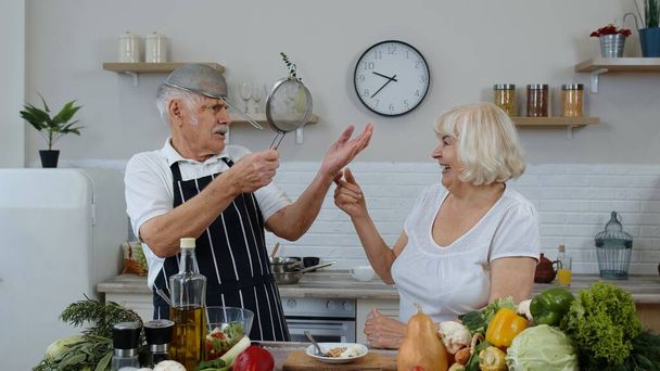 シニアの女と男がストレーナーと面白いダンスをする。台所で一緒に料理をしながら踊る - 写真・画像