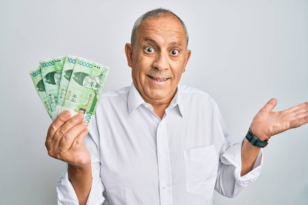 Handsome senior man holding 10000 south korean won banknotes celebrating achievement with happy smile and winner expression with raised hand  - Fotoğraf, Görsel