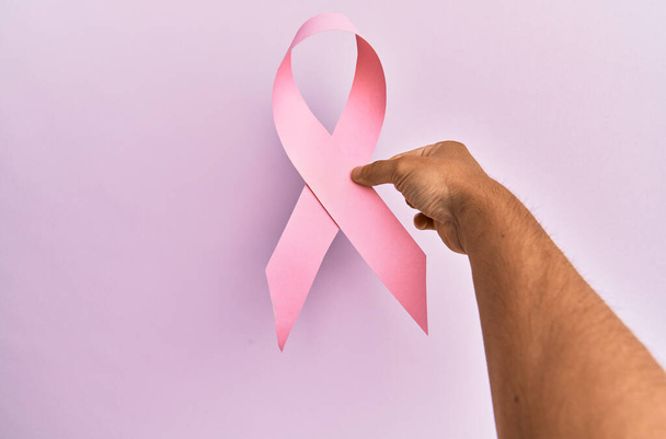 Mano de joven hispano sosteniendo cinta rosa de cáncer de mama sobre fondo aislado. - Foto, imagen