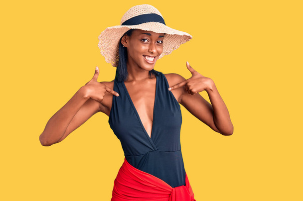 Joven mujer afroamericana vistiendo traje de baño y sombrero de verano mirando confiado con sonrisa en la cara, señalándose con los dedos orgullosos y felices.  - Foto, Imagen