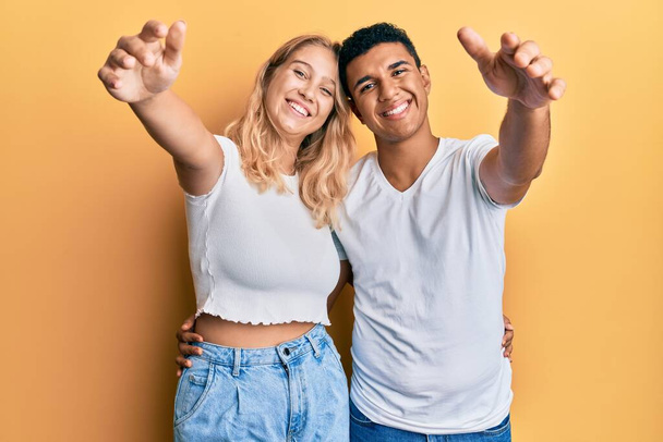 若い異人種間のカップルは抱擁のために開いて腕で笑みを浮かべてカメラを見てカジュアルな白いTシャツを着て。幸せを受け入れる陽気な表情.  - 写真・画像