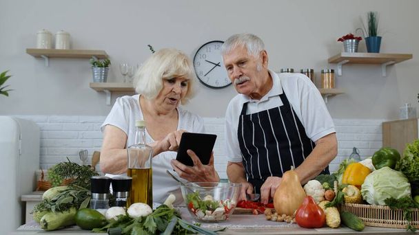 Vegan senior grandparents looking for a culinary recipe online on digital tablet, cooking salad - Foto, imagen