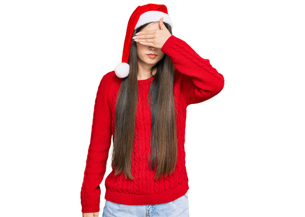 Joven mujer china con sombrero de navidad cubriendo los ojos con la mano, luciendo seria y triste. concepto de ocultamiento y rechazo sin visión  - Foto, Imagen