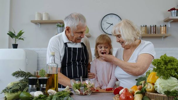 Elderly grandparents in kitchen feeding grandchild girl with chopped red pepper. Vegetarian diet - Foto, imagen