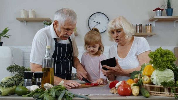 Ηλικιωμένοι παππούδες ζευγάρι με ψηφιακή ταμπλέτα και εγγονή κοπή λαχανικών στην κουζίνα - Φωτογραφία, εικόνα