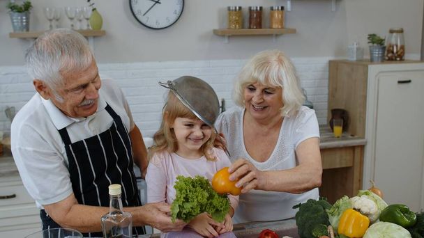 Boldog vegán idős pár táncol unokája gyermek főzés közben zöldségek a konyhában - Fotó, kép