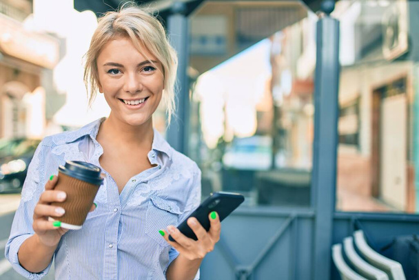 Joven mujer rubia sonriendo feliz usando el teléfono inteligente y bebiendo llevar café en la ciudad. - Foto, imagen