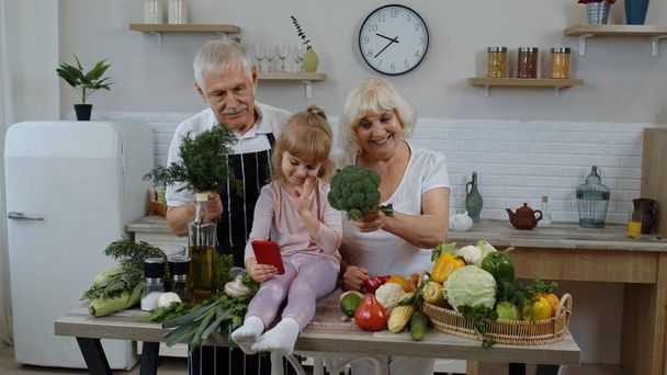 Blogcu kız cep telefonuyla fotoğraf çekiyor. Büyük anne ve büyükbabasıyla mutfakta sebzeli. - Fotoğraf, Görsel