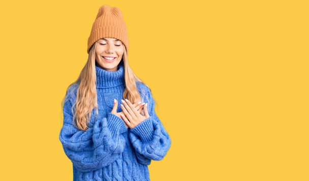 ブロンドの髪をした美しい白人女性は、ウールのセーターと冬の帽子を着て、目を閉じて胸に手で笑顔と顔に感謝のジェスチャー。健康の概念.  - 写真・画像