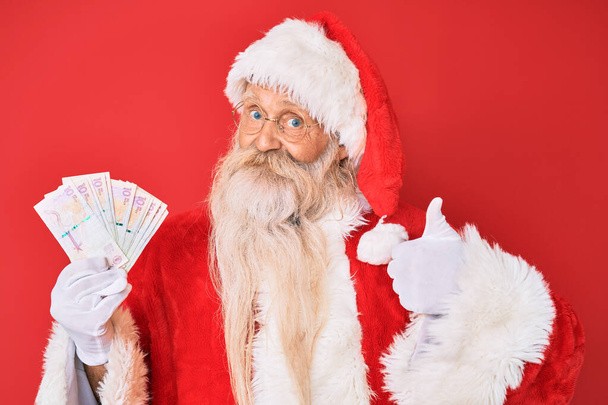 Staruszek z siwymi włosami i długą brodą w kostiumie Świętego Mikołaja trzymający kolumbijskie peso uśmiechnięte szczęśliwe i pozytywne, kciuk do góry robi doskonałe i znak aprobaty  - Zdjęcie, obraz
