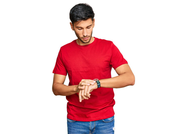 Молодой красивый мужчина в обычной красной футболке проверяя время на наручных часах, расслабленной и уверенной  - Фото, изображение
