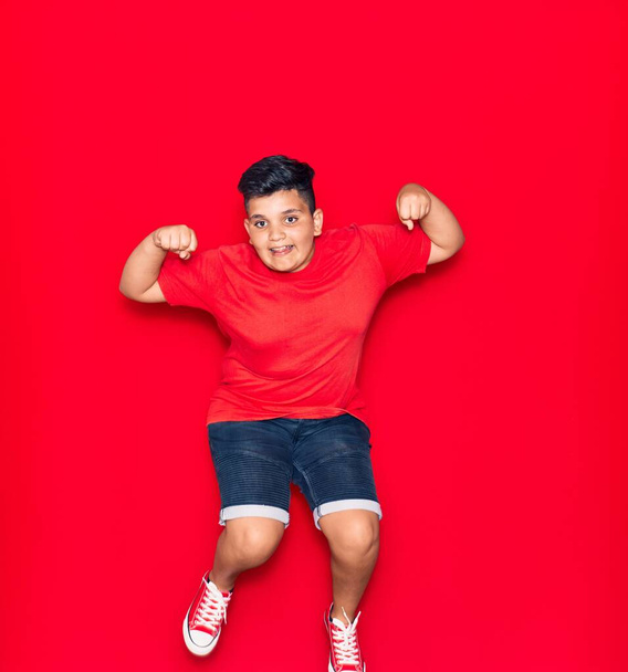 Adorable kid wearing casual clothes smiling happy. Jumping with smile on face doing strong gesture with arms over isolated red background - Valokuva, kuva