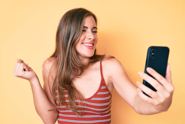 Beautiful young caucasian woman taking a selfie photo with smartphone screaming proud, celebrating victory and success very excited with raised arm  - Foto, Bild