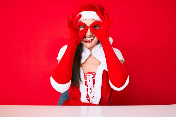 Young caucasian woman wearing santa claus costume doing ok gesture like binoculars sticking tongue out, eyes looking through fingers. crazy expression.  - Foto, Bild