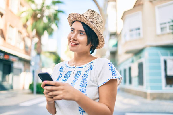 jong latin toeristisch meisje op vakantie glimlachend gelukkig met behulp van smartphone in de stad. - Foto, afbeelding