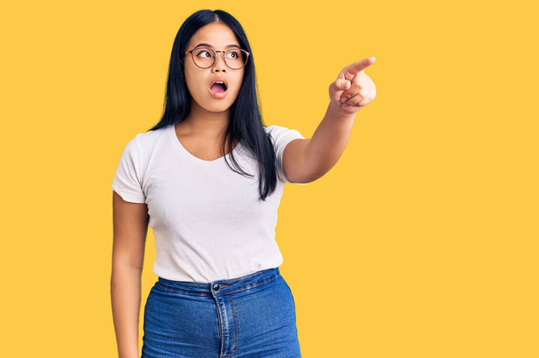 Young beautiful asian girl wearing casual clothes and glasses pointing with finger surprised ahead, open mouth amazed expression, something on the front  - 写真・画像