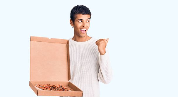 Молодий афроамериканський чоловік тримає коробку доставки піци вказуючи великий палець в сторону посміхаючись щасливим з відкритим ротом
  - Фото, зображення